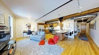 This thoughtfully designed single-floor space blends industrial character with modern comfort in an open-concept layout. Exposed wooden ceiling beams and black track lighting run the length of the unit, lending a loft-like aesthetic that ties the living, dining, and kitchen areas together seamlessly. Warm hardwood flooring flows throughout, enhancing the sense of openness and continuity.

The kitchen is well-appointed with stainless steel appliances, including a full-size refrigerator, stove, and built-in microwave, all framed by rich wood cabinetry. A curved kitchen island with a light countertop provides bar-style seating, making it ideal for casual dining or entertaining. Pendant lighting above the island adds warmth and focused illumination.

The living area is anchored by a plush gray sectional sofa and a patterned area rug, complemented by a pair of bold red accent chairs that bring a vibrant, contemporary energy to the space. A dedicated workspace with a desk and office chair is neatly positioned near the perimeter, offering a functional area for work or study. Wall art adds a curated, artistic touch throughout.

A separate dining nook with a table and seating rounds out the layout, making this space as practical as it is visually striking.