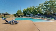 Located at 1360 Park Ashwood Dr in Saint Charles, Missouri, this outdoor amenity area serves as a vibrant recreational hub designed for relaxation and community enjoyment. At its centerpiece is a generously sized freeform swimming pool with a curvilinear design, featuring a shallow entry zone and a classic brick coping border that lends the space a polished, resort-style aesthetic. The pool's clear turquoise water is complemented by a spacious surrounding deck finished in a warm tan aggregate surface, providing ample room for lounging and socializing. Numerous green chaise lounge chairs are arranged throughout the deck, accompanied by coordinating blue market umbrellas that offer shade on sunny days. A covered pergola structure anchors one side of the deck, providing an additional shaded gathering spot. Adjacent to the pool area, a colorful children's playground adds a family-friendly element to the amenity space. The entire area is enclosed by a secure perimeter fence, ensuring privacy and safety. A single-story clubhouse building with a pitched roof and large windows borders the pool, offering convenient indoor-outdoor access. Mature trees lining the perimeter provide natural shade and a lush green backdrop, completing this well-appointed outdoor retreat.