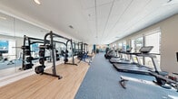 This single-story fitness center is thoughtfully designed to accommodate a wide range of training needs across several distinct zones. The main workout area is generously proportioned, with a drop-panel ceiling, recessed lighting, and large windows along one wall that invite abundant natural light and views of the surrounding landscape. The flooring transitions strategically between light hardwood-style planks in the free weights section and blue carpet beneath the cardio equipment, providing both functional and visual differentiation between zones.

The cardio section features a row of treadmills and stair climbers positioned to face the windows, while the strength training area houses weight racks, barbells, cable machines, and adjustable benches backed by full-length mirrors. A dedicated functional training zone includes a multi-station rig, kettlebells, medicine balls, sandbags, and rubber flooring for high-intensity workouts. A large motivational mural adds visual energy to the interior.

A separate studio room offers hardwood-style flooring, wall-to-wall mirrors, mounted monitors, and direct access to the outdoors through glass doors. The exterior features manicured artificial turf, palm trees, iron fencing, and a waterfront backdrop, creating an inviting setting that extends the fitness experience beyond the walls.