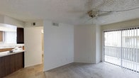 This single-floor residential space offers a clean, functional layout designed for comfortable everyday living. The main living area is finished with wall-to-wall carpeting and features a ceiling fan overhead, providing both comfort and airflow throughout the space. Smooth white walls and a textured ceiling give the room a bright, neutral aesthetic that allows for easy personalization. A sliding glass door opens directly to a private balcony, inviting natural light into the interior and offering a seamless connection to the outdoors. An HVAC vent is integrated into the wall, ensuring efficient climate control year-round. Adjacent to the main living area, a transition to tile flooring signals a move toward the kitchen or utility spaces, visible through an open passageway. The open sightlines between rooms enhance the overall sense of space and flow throughout the unit. The minimalist, move-in-ready design provides a versatile canvas suitable for a range of furnishing styles and living arrangements.