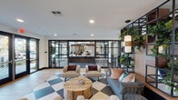 This thoughtfully designed single-floor amenity space offers a dynamic blend of social, recreational, and functional zones seamlessly connected through cohesive interior design. The layout transitions naturally between areas, beginning with a welcoming lounge featuring a striking black-and-white checkered tile floor anchored by plush seating arrangements, a rustic wood coffee table, and colorful accent pillows. Floor-to-ceiling black-framed glass partitions enclose a bar area with a patterned tile backsplash, dark wood cabinetry, and bar stool seating, maintaining visual connectivity throughout the space.

An open great room commands attention with soaring double-height ceilings, oversized windows that flood the interior with natural light, and sculptural pendant light fixtures. This area accommodates multiple seating configurations alongside a pool table and a dedicated media wall featuring built-in shelving, integrated greenery, and dual flat-screen televisions. Light wood plank flooring extends through the co-working and game areas, providing warmth and continuity.

A well-appointed kitchenette and mailroom corridor showcase rich walnut cabinetry, deep green subway tile, open shelving with lush plants, and an organized package and mailbox wall. Black metal open shelving units adorned with trailing plants serve as decorative dividers throughout, reinforcing the space's urban-botanical aesthetic.