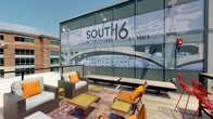 The South 16 at The Bridges Roof Deck is a well-appointed outdoor amenity space that combines urban design with relaxed comfort. Accessed via a carpeted interior corridor accented with teal walls and warm lighting, the transition from the building's interior to the deck is seamless and welcoming. A dedicated entryway with Adirondack chairs provides a casual waiting area before stepping out onto the expansive open-air deck.

The deck itself features a wood-plank surface and is outfitted with curated lounge furniture, including wicker chairs with plush white and orange cushions, leather-accented side chairs, and gold-toned accent tables arranged over vibrant patterned area rugs. The eclectic yet cohesive furnishings create a comfortable and inviting atmosphere ideal for relaxation and socializing.

A defining architectural feature of the deck is the large-scale branded mural depicting a bridge, a nod to the property's namesake and the surrounding area's identity. Industrial-style wall sconces accent the mural wall, adding both function and visual character. The deck is enclosed by sleek railing systems that maintain open sightlines to the surrounding cityscape, neighboring structures, and lush green hillsides visible in the distance. Under open skies, this rooftop retreat offers a dynamic and engaging outdoor experience.