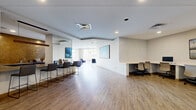 This cyber cafe and clubhouse spans a generously sized single floor, offering a multifunctional community space designed with a clean, contemporary aesthetic. The layout is thoughtfully organized into distinct zones that encourage both productivity and relaxation. Wood-look flooring flows throughout the majority of the space, complemented by light-toned walls and recessed lighting that creates a bright, welcoming atmosphere.

The cyber cafe area features a built-in workstation desk with ergonomic seating, providing a dedicated space for computer use, alongside a bar-height counter with sleek stools suited for casual work or socializing. Large wall-mounted displays and curated artwork are positioned throughout, adding visual interest to the open layout.

The clubhouse lounge area is anchored by a striking floor-to-ceiling tiled accent wall housing a linear fireplace, with a large mounted television above. Comfortable seating, including a neutral sectional sofa and accent chairs, creates an inviting gathering space. Expansive floor-to-ceiling windows and glass doors flood the interior with natural light and provide direct access to an outdoor patio. The entry area features a decorative partition screen and glass-panel doors, offering a polished first impression upon arrival.