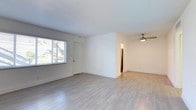 This single-floor space is defined by a clean, minimalist aesthetic carried consistently throughout. Light wood-look flooring runs seamlessly across the living areas, complementing the crisp white walls and bright, airy atmosphere created by natural light filtering through horizontal-slatted blinds.

The open living area is generously proportioned, offering flexible layout options. A ceiling fan with a light fixture provides both airflow and ambient lighting, while a central HVAC vent in the ceiling ensures climate control. The entry area features a front door flanked by a large window that frames outdoor tropical greenery, reinforcing the bright, welcoming character of the space.

A hallway connects the living area to additional rooms, one of which features a louvered closet door visible from the main space. A flush-mount ceiling light illuminates the hallway corridor.

The kitchen is efficiently designed in a galley layout, outfitted with white cabinetry featuring modern bar-pull hardware, granite countertops, a stainless steel electric range, an under-cabinet range hood, and a single-basin sink beneath a window. Track lighting overhead provides focused task illumination. Throughout, the space maintains a cohesive, updated, and move-in-ready presentation.