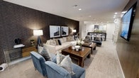 This single-floor space exudes a polished, contemporary elegance that balances comfort with refined design. The lounge area serves as the centerpiece, anchored by a striking dark geometric-patterned accent wall that adds depth and drama to the room. A large, cream-toned sofa is paired with plush navy blue accent chairs, all accessorized with decorative pillows in coordinating geometric prints, creating a cohesive and inviting seating arrangement. Rich, warm-toned wooden coffee tables anchor the seating cluster, styled with tasteful décor including floral arrangements and sculptural accents. Recessed lighting throughout the ceiling provides a clean, modern look while illuminating the space evenly, complemented by elegant floor lamps that add warmth. The light-toned flooring extends seamlessly throughout, lending an open and airy feel to the interior. Framed artwork adorns the accent wall, reinforcing the space's curated aesthetic. Adjacent to the lounge, a dining area is visible in the background, featuring dark bistro-style chairs arranged around tables, suggesting a versatile environment well-suited for both casual lounging and social dining. The overall ambiance is sophisticated, welcoming, and thoughtfully appointed.