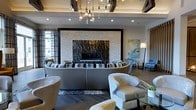This single-floor space showcases a thoughtfully designed interior that blends contemporary elegance with warm, inviting elements. The focal point of the room is a dramatic floor-to-ceiling stone fireplace, featuring a striking combination of stacked natural stone surround and a dark, richly textured stone hearth panel that commands immediate attention upon entry. A curved, wraparound sectional sofa with a bold striped pattern anchors the central seating area, complemented by a variety of plush accent chairs in neutral and blue tones that invite relaxed conversation. Overhead, an artistic chandelier with exposed Edison bulbs suspended at varying heights provides a striking visual element, while recessed lighting ensures consistent, warm illumination throughout the space. Dark wood beam detailing near the ceiling adds a rustic contrast to the otherwise modern aesthetic. Rich hardwood flooring extends across the room, grounding the space with warmth and continuity. Patterned area rugs define seating zones, while large windows allow natural light to filter in. Carefully curated artwork and lush greenery complete the sophisticated, well-appointed ambiance of this stylish communal lounge.