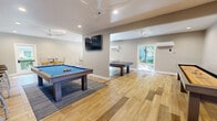 This single-floor game room is a dedicated entertainment space designed to accommodate a wide variety of recreational activities. The layout is open and generously proportioned, offering ample room for multiple games and social gatherings simultaneously. Light wood-look flooring spans the entire space, lending a clean, contemporary aesthetic that pairs well with the neutral gray walls and white ceilings.

At the center of the room, a pool table with a vibrant blue felt surface serves as the focal point, positioned atop a patterned area rug. A wall-mounted flat-screen TV is situated nearby, ideal for watching sports or entertainment between games. A ping-pong table occupies its own dedicated zone, while a shuffleboard table and foosball table round out the impressive lineup of gaming options.

Recessed lighting provides bright, even illumination throughout, supplemented by multiple ceiling fans that promote airflow and comfort. A mini-split HVAC unit ensures climate control year-round. A seating area with bar-height tables and chairs offers a convenient spot to relax, socialize, or spectate. A sliding glass door at one end of the room allows natural light to pour in and provides access to an outdoor area. The overall ambiance is modern, inviting, and purpose-built for leisure and entertainment.