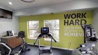 This single-floor fitness center is thoughtfully designed to support a well-rounded workout experience. The main exercise area is defined by a bold, chartreuse accent wall emblazoned with the motivational phrase "Work Hard and Be Proud of What You Achieve," setting an energetic tone throughout the space. The room is outfitted with a range of cardio equipment, including a Landice treadmill, an elliptical trainer, and a recumbent stationary bike, providing users with versatile options for cardiovascular exercise. A dedicated strength-training area houses a multi-functional cable machine with an adjustable bench, accommodating a variety of resistance exercises. Durable dark rubberized flooring covers the exercise area, providing both safety and comfort underfoot. Natural light streams in through multiple double-hung windows that offer views of the surrounding outdoor landscape, while recessed and overhead lighting ensure the space is well-lit at all times. A wall-mounted television adds entertainment value during workouts. A small refreshment station with a coffee maker is conveniently situated in the corner. Elegant white French doors connect the fitness room to a bright, furnished lounge area, creating a seamless flow between activity and relaxation.