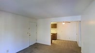 This single-floor space features a straightforward, open layout that connects the main living area to the kitchen. The living area is finished with wall-to-wall carpet in a neutral tone, providing a simple and practical foundation. White walls throughout give the space a clean, understated appearance, and a textured ceiling adds a subtle architectural detail overhead.

A standard entry door with a brass handle is positioned along one wall of the living area, providing direct access to the unit. The space opens into the kitchen area, which is visible through a wide pass-through opening. The kitchen is equipped with medium-toned wood cabinetry — both upper and lower units — along with a sink and countertop workspace. Dark-toned flooring in the kitchen contrasts with the carpeted living area, clearly delineating the two spaces.

A ceiling fan with a built-in light fixture is installed in the kitchen or adjoining area, offering both ventilation and ambient lighting. The overall layout is compact and functional, with a no-frills design that prioritizes practicality. The neutral color palette and simple finishes create a cohesive, easily adaptable environment throughout the space.