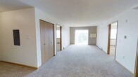 This single-floor space features an open, airy layout with neutral white walls and soft, plush wall-to-wall carpeting throughout, creating a clean and comfortable atmosphere. Natural light floods the interior through a large sliding glass door at the far end of the room, which opens to an outdoor balcony or patio area, offering views of the surrounding environment. The generous proportions of the main living area provide a versatile open floor plan adaptable to various furniture arrangements. A set of wooden bi-fold closet doors along one wall offers substantial built-in storage, adding both functionality and a warm wood-tone contrast to the otherwise neutral palette. An adjacent room or hallway is visible through an open doorway, suggesting additional living or bedroom space beyond the main area. Baseboard heating units run along the perimeter walls, indicating a practical and efficient heating system. The overall ambiance is clean, bright, and move-in ready, characterized by its simple, understated finishes and an abundance of natural light.