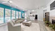 Located in Gulf Shores, Alabama, this single-floor space offers a well-designed, open layout encompassing a spacious living room and a fully equipped kitchen. The living room is a standout area, featuring a classic fireplace as a focal point, complemented by a wall-mounted television above. Crown molding lines the ceilings throughout, adding a refined architectural touch, while multiple ceiling fans ensure comfortable airflow. Light-toned hardwood-style flooring runs seamlessly across the entire space, enhancing the bright, airy feel. A bold teal accent wall enlivens the living area, contrasting beautifully with the crisp white walls and trim. A large bank of windows along one wall floods the room with natural light and offers views of the outdoor surroundings, including a glimpse of a pool area. The kitchen is thoughtfully designed with white cabinetry, a neutral tile backsplash, marble-style countertops, and black appliances including a refrigerator and range. A peninsula breakfast bar provides additional prep and seating space, creating a natural connection between the kitchen and the living area. Recessed lighting and a textured ceiling complete the interior. The overall ambiance is coastal, clean, and inviting — an ideal setting for relaxed Gulf Coast living.