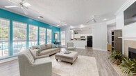 Located in Gulf Shores, Alabama, this single-floor space offers a well-designed, open layout encompassing a spacious living room and a fully equipped kitchen. The living room is a standout area, featuring a classic fireplace as a focal point, complemented by a wall-mounted television above. Crown molding lines the ceilings throughout, adding a refined architectural touch, while multiple ceiling fans ensure comfortable airflow. Light-toned hardwood-style flooring runs seamlessly across the entire space, enhancing the bright, airy feel. A bold teal accent wall enlivens the living area, contrasting beautifully with the crisp white walls and trim. A large bank of windows along one wall floods the room with natural light and offers views of the outdoor surroundings, including a glimpse of a pool area. The kitchen is thoughtfully designed with white cabinetry, a neutral tile backsplash, marble-style countertops, and black appliances including a refrigerator and range. A peninsula breakfast bar provides additional prep and seating space, creating a natural connection between the kitchen and the living area. Recessed lighting and a textured ceiling complete the interior. The overall ambiance is coastal, clean, and inviting — an ideal setting for relaxed Gulf Coast living.