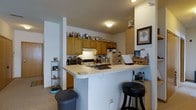 This single-floor residence centers around a compact yet well-organized kitchen that serves as a focal point of the living space. The kitchen features medium-toned oak cabinetry that extends to the ceiling, providing generous storage capacity. Upper cabinets line the walls, offering ample room for pantry items and kitchenware, with additional storage utilized along the tops of the cabinets.

A white peninsula counter extends outward from the kitchen, creating a natural divider between the cooking area and the adjoining living space. This peninsula doubles as a casual dining and workspace, accommodated by a black bar stool tucked underneath. The countertop provides a practical surface for everyday tasks and informal dining.

The kitchen is equipped with standard appliances, including a range with an overhead ventilation hood and a refrigerator. A microwave and countertop appliances round out the cooking setup. Flush-mount ceiling lighting provides consistent illumination throughout the space.

A small shelving unit in the corner adds supplemental storage and display space. The overall ambiance is practical and lived-in, with neutral walls and light-toned flooring contributing to a cohesive, functional interior.