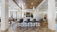 This generously sized single-floor amenity space offers a sophisticated, multi-functional layout designed for both socializing and focused activity. The interior immediately impresses with its striking geometric-patterned wood-look tile flooring, which transitions seamlessly into plush neutral carpeting in the lounge and game areas. A prominent central bar anchors the main gathering zone, featuring a long white quartz countertop lined with leather-upholstered barstools on brass-toned frames, illuminated by a series of white conical pendant lights suspended from a warm wood-paneled ceiling inset.

Adjacent to the bar, a dedicated coffee and refreshment station includes a commercial-grade coffee machine, sink, and under-counter refrigeration. The lounge area offers generous seating arrangements centered around a wood-accented fireplace surround with a wall-mounted display, flanked by floor-to-ceiling windows that invite abundant natural light. A game room section features a shuffleboard table set against large windows with linen drapery. Private conference or meeting rooms with sliding glass-paneled doors provide quieter, enclosed settings. Throughout, coffered ceilings, white structural columns, and recessed lighting contribute to a polished, cohesive ambiance.