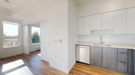 This single-floor unit showcases a clean, contemporary design characterized by crisp white walls, warm-toned hardwood flooring, and an efficient layout. The kitchen is a standout feature, outfitted with a full suite of stainless steel appliances including a refrigerator, electric range with overhead ventilation, and dishwasher. Flat-panel white upper cabinetry reaches toward the ceiling, maximizing storage, while the lower cabinets feature a contrasting wood-grain finish. A white subway tile backsplash and light countertops complete the polished aesthetic.

A partial wall separates the kitchen from the living area, which benefits from generous natural light streaming through large windows that frame views of surrounding treetops and the neighborhood below. Baseboard heating runs along the exterior walls, ensuring year-round comfort.

The bedroom is a calm, sunlit space with continued hardwood flooring and an open closet with a rod for hanging storage. The bathroom offers a distinctive touch with bold geometric patterned floor tile in black and white, paired with a sleek dark-toned vanity, white integrated sink, frameless mirror, and clean white walls, creating a stylish yet functional space throughout.