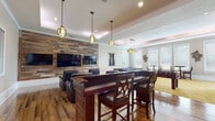 Located at 906 Oasis Palm Circle in Ocoee, Florida, this single-story community clubhouse is designed for entertainment and social gathering. The space showcases a compelling blend of rustic and contemporary design, anchored by rich hardwood flooring, tray ceilings with crown molding, and distinctive lantern-style pendant lighting throughout.

The centerpiece of the clubhouse is a spacious media lounge featuring a dramatic reclaimed wood accent wall that spans the length of the room, upon which multiple flat-screen televisions are mounted. Plush leather recliner chairs are arranged for optimal viewing comfort. Adjacent to the media area is a bar-height seating section with a dark wood communal table and upholstered bar stools, ideal for casual socializing.

A dedicated game room area offers a billiards table, foosball table, and classic arcade and pinball machines, providing a variety of entertainment options. A separate carpeted lounge area — finished with a bold, decorative gold-toned carpet — features bistro-style tables with leather chairs positioned beneath large windows that invite natural light and views of the surrounding greenery.

The overall ambiance is warm and inviting, combining functional design with thoughtful décor, including vibrant wall art. The space is well-lit with recessed lighting supplementing the decorative pendants, creating a welcoming atmosphere for residents to gather and relax.