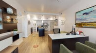 The Devon at University Place is a single-floor space that functions as both a model unit and leasing office, offering prospective residents a comprehensive look at the community's design and finishes. The layout flows openly between a leasing reception area and a fully appointed model unit, all unified by warm-toned luxury vinyl plank flooring throughout the common and living areas.

The leasing office features a dedicated workspace with a dark-finished desk, ergonomic seating, and a welcoming guest area with green upholstered chairs, framed artwork, and large windows that invite natural light. A ceiling fan overhead contributes to an airy, comfortable atmosphere.

The model kitchen is a standout feature, anchored by a bold navy island with a light countertop and bar-height seating. White upper cabinetry contrasts with dark lower cabinets, and stainless steel appliances complete the modern aesthetic. Open shelving adds functional character to the space.

The bedroom is carpeted and comfortably scaled, featuring a ceiling fan, natural light from a window with exterior views, and warm wood-toned furnishings. The bathroom is cleanly designed with a tub-shower combination, white subway tile surround, dark vanity cabinetry, and light wood-look flooring that ties cohesively to the rest of the unit.