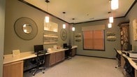 This single-floor space serves as a thoughtfully designed business center, offering a functional and welcoming environment for focused work. The room features a long, built-in wooden workstation that spans the length of one wall, providing ample desk space accommodating multiple computer workstations with ergonomic office chairs. Warm-toned cabinetry beneath the desk offers generous storage while maintaining a clean, organized appearance.

Overhead, a series of pendant lights with frosted glass shades hang from the ceiling, casting a warm, even glow throughout the room. The walls are painted in a muted sage green, complemented by rich dark crown molding that adds a refined architectural detail. Decorative wall art, including woven circular accents, textured panels, and nature-inspired pieces, lends the space a sophisticated yet approachable character.

Natural light filters into the room through a window dressed with warm-toned wooden blinds, softening the overall ambiance. The carpeted flooring in a neutral tone adds comfort underfoot and contributes to the room's quiet, productive atmosphere. Together, the design elements balance practicality with style, creating a space that is both highly functional and visually inviting.