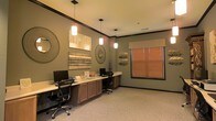 This single-floor space serves as a thoughtfully designed business center, offering a functional and welcoming environment for focused work. The room features a long, built-in wooden workstation that spans the length of one wall, providing ample desk space accommodating multiple computer workstations with ergonomic office chairs. Warm-toned cabinetry beneath the desk offers generous storage while maintaining a clean, organized appearance.

Overhead, a series of pendant lights with frosted glass shades hang from the ceiling, casting a warm, even glow throughout the room. The walls are painted in a muted sage green, complemented by rich dark crown molding that adds a refined architectural detail. Decorative wall art, including woven circular accents, textured panels, and nature-inspired pieces, lends the space a sophisticated yet approachable character.

Natural light filters into the room through a window dressed with warm-toned wooden blinds, softening the overall ambiance. The carpeted flooring in a neutral tone adds comfort underfoot and contributes to the room's quiet, productive atmosphere. Together, the design elements balance practicality with style, creating a space that is both highly functional and visually inviting.