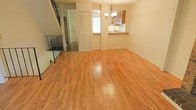 This three-floor home offers a well-organized layout designed for comfortable everyday living. The main living area is a standout feature, showcasing gleaming hardwood floors that extend throughout the space, lending warmth and a sense of openness. A staircase with a classic wrought-iron railing connects the levels, adding an elegant architectural touch while maximizing the vertical flow of the home. Natural light filters in through a window, brightening the living area and highlighting the clean, neutral wall tones that provide a versatile backdrop for any style of décor. A convenient pass-through window connects the living area to the kitchen, which features wood-toned cabinetry and a countertop bar — ideal for casual dining or entertaining. The kitchen's practical design makes efficient use of the space without sacrificing style. A closet with louvered bi-fold doors provides ample storage directly off the living area, while an additional door leads to further spaces within the home. The multi-level layout offers distinct zones for living, sleeping, and additional functional use, making this a highly adaptable home suited to a variety of lifestyles. The overall ambiance is bright, clean, and move-in ready.