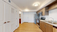 Located at 10503 E Burnside St in Portland, OR, this two-bedroom, one-bathroom unit offers a well-designed single-floor layout that maximizes functionality. The open-plan kitchen and living area form the heart of the space, where light wood-tone laminate flooring creates a clean, modern aesthetic. The kitchen is outfitted with stainless steel appliances—including a range, microwave, refrigerator, and dishwasher—complemented by warm walnut-toned cabinetry, white countertops, and a double basin sink. Flush-mount ceiling lighting keeps the area bright and well-illuminated throughout. The living area transitions seamlessly from the kitchen, featuring plush carpeting and a window with vertical blinds that allows natural light to filter in. Both bedrooms are carpeted and finished with neutral white walls; the primary bedroom includes a walk-in closet with wire shelving for organized storage. The bathroom features a wood-toned vanity with a white countertop, integrated sink, and a well-lit mirror above. Consistent design elements—matching cabinetry, light flooring, and clean white walls—give the unit a cohesive, contemporary feel throughout.