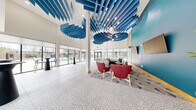 This contemporary amenity lounge is a generously proportioned single-floor space designed to impress and inspire. The most striking architectural feature is the dramatic double-height ceiling adorned with cascading blue acoustic baffle panels, which serve both a functional and artistic purpose, creating a bold visual centerpiece throughout the main gathering area. Globe pendant lights complement the baffles, adding warmth to the soaring overhead expanse.

The flooring throughout is a sophisticated terrazzo with a crackle-pattern design in neutral tones, lending texture and continuity across all areas. Floor-to-ceiling windows and sliding glass doors line the perimeter, inviting abundant natural light and offering open views of the surrounding outdoor spaces, including a pool area visible from the lounge.

The main lounge area features a rich mix of seating arrangements, including curved red accent chairs, a neutral sectional sofa paired with a high bar table and yellow stools, and cocktail-height tables with sleek black pedestals. A bold teal accent wall serves as a dramatic backdrop for dual mounted flat-screen televisions, anchoring the social hub of the space.

A curved reception counter with warm wood slat detailing and blue upholstered barstools adds a polished, hospitality-inspired touch. A separate, glass-partitioned meeting room provides a more intimate setting with a round table and yellow stools, equipped with a wall-mounted display screen.
