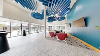This contemporary amenity lounge is a generously proportioned single-floor space designed to impress and inspire. The most striking architectural feature is the dramatic double-height ceiling adorned with cascading blue acoustic baffle panels, which serve both a functional and artistic purpose, creating a bold visual centerpiece throughout the main gathering area. Globe pendant lights complement the baffles, adding warmth to the soaring overhead expanse.

The flooring throughout is a sophisticated terrazzo with a crackle-pattern design in neutral tones, lending texture and continuity across all areas. Floor-to-ceiling windows and sliding glass doors line the perimeter, inviting abundant natural light and offering open views of the surrounding outdoor spaces, including a pool area visible from the lounge.

The main lounge area features a rich mix of seating arrangements, including curved red accent chairs, a neutral sectional sofa paired with a high bar table and yellow stools, and cocktail-height tables with sleek black pedestals. A bold teal accent wall serves as a dramatic backdrop for dual mounted flat-screen televisions, anchoring the social hub of the space.

A curved reception counter with warm wood slat detailing and blue upholstered barstools adds a polished, hospitality-inspired touch. A separate, glass-partitioned meeting room provides a more intimate setting with a round table and yellow stools, equipped with a wall-mounted display screen.