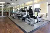 This single-floor fitness and recreation facility is thoughtfully designed to offer a comprehensive wellness and leisure experience. The space is divided into distinct yet visually connected zones, separated by floor-to-ceiling glass partition walls and sliding glass doors that allow natural light to flow freely throughout.

The primary cardio area features a row of modern treadmills and elliptical machines by TRUE Fitness, positioned in front of large windows that frame lush, wooded hillside views and an outdoor terrace with seating. Ceiling fans overhead and a drop-tile ceiling with skylights keep the environment bright and comfortable. Wood-look flooring runs through the main exercise zones, while rubberized matting anchors the cardio equipment.

An adjacent multi-use fitness room offers additional exercise equipment, including a recumbent bike and open floor space suitable for stretching or group workouts. A separate strength training area is visible through the glass walls, housing additional fitness machines.

Completing the facility is a stylish billiards and lounge room, featuring a full-size pool table, high-top bar seating with teal upholstered stools, and a decorative lattice-patterned accent wall. The cohesive design and open sightlines throughout the space create an inviting, social atmosphere ideal for both active and recreational use.