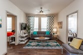 This single-story unit offers a thoughtfully designed layout with consistent finishes and a contemporary aesthetic carried throughout each room. Rich hardwood-style flooring flows seamlessly across all living spaces, anchoring the design with warmth and cohesion.

The living room serves as the social hub of the unit, featuring large windows dressed with patterned drapes that allow generous natural light to fill the space. A ceiling fan overhead and a neutral color palette create a comfortable, airy atmosphere well-suited for both relaxing and entertaining.

The kitchen is a standout feature, outfitted with dark cabinetry, granite-style countertops, and a full suite of stainless steel appliances including a refrigerator, range, and over-the-range microwave. A peninsula with bar seating connects the kitchen to the entry corridor, providing a casual dining option and enhancing the open feel of the space.

Both bedrooms are comfortably proportioned, each with a ceiling fan, a window with natural light, and sufficient space for a full bedroom setup including a bed with under-drawer storage and a dedicated workspace.

The two full bathrooms are consistently finished, each featuring a tub-shower combination with gray subway tile surround, dark vanity cabinetry, and granite-style countertops, offering a clean and polished look.