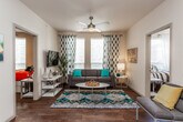 This single-story unit offers a thoughtfully designed layout with consistent finishes and a contemporary aesthetic carried throughout each room. Rich hardwood-style flooring flows seamlessly across all living spaces, anchoring the design with warmth and cohesion.

The living room serves as the social hub of the unit, featuring large windows dressed with patterned drapes that allow generous natural light to fill the space. A ceiling fan overhead and a neutral color palette create a comfortable, airy atmosphere well-suited for both relaxing and entertaining.

The kitchen is a standout feature, outfitted with dark cabinetry, granite-style countertops, and a full suite of stainless steel appliances including a refrigerator, range, and over-the-range microwave. A peninsula with bar seating connects the kitchen to the entry corridor, providing a casual dining option and enhancing the open feel of the space.

Both bedrooms are comfortably proportioned, each with a ceiling fan, a window with natural light, and sufficient space for a full bedroom setup including a bed with under-drawer storage and a dedicated workspace.

The two full bathrooms are consistently finished, each featuring a tub-shower combination with gray subway tile surround, dark vanity cabinetry, and granite-style countertops, offering a clean and polished look.