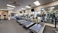 Located at 475 W Enterprise Dr in Mount Prospect, Illinois, this fitness center offers a comprehensive single-floor facility designed to support a wide range of workout routines. The main exercise floor features durable rubber flooring and is lined with floor-to-ceiling mirrors that enhance the sense of openness throughout the space. Bright recessed panel lighting and ceiling fans ensure the area remains well-lit and comfortable during use.

The cardio section is equipped with a row of treadmills, ellipticals, and steppers, positioned near windows that allow natural light to filter in and provide views of the surrounding outdoor environment. Wall-mounted televisions are strategically placed throughout for entertainment during workouts. The strength training area houses a comprehensive selection of selectorized machines, a well-stocked dumbbell rack with a flat bench, and additional cable and resistance equipment.

A dedicated training zone features hardwood-style flooring and is stocked with medicine balls, stability balls, and resistance tools, creating an ideal space for functional and group-style training. A Peloton bike station adds a modern touch for cycling workouts. A water fountain is conveniently accessible within the facility. The overall ambiance is clean, functional, and organized, making this fitness center a well-rounded destination for health and wellness.