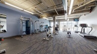 Located at 320 W Broadway in Monona, Wisconsin, this fitness center offers a well-appointed, single-floor workout facility designed to support a comprehensive exercise regimen. The space features an industrial-inspired aesthetic, with exposed concrete ceilings, visible ductwork and piping, and structural columns that lend the room a raw, urban character. Durable, patterned carpet tile flooring covers the main workout areas, while rubber flooring provides added traction in the free weights zone.

The facility is divided into distinct workout areas. The cardio section is equipped with treadmills, stationary bikes, elliptical machines, and rowing equipment, positioned near large windows that allow natural light to fill the space and offer views of the surrounding outdoor environment. A dedicated strength training area houses cable machines, functional trainers, and multi-use weight stations. A separate free weights zone features a full dumbbell rack, barbells, weight plates, squat racks, benches, and medicine balls, complemented by wall-mounted mirrors for form monitoring.

Overhead linear fluorescent lighting ensures bright, even illumination throughout. A wall-mounted television provides entertainment during workouts. The open layout and generous ceiling height create an airy, functional environment well-suited for a variety of fitness activities.