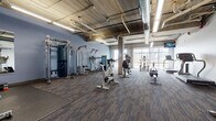 Located at 320 W Broadway in Monona, Wisconsin, this fitness center offers a well-appointed, single-floor workout facility designed to support a comprehensive exercise regimen. The space features an industrial-inspired aesthetic, with exposed concrete ceilings, visible ductwork and piping, and structural columns that lend the room a raw, urban character. Durable, patterned carpet tile flooring covers the main workout areas, while rubber flooring provides added traction in the free weights zone.

The facility is divided into distinct workout areas. The cardio section is equipped with treadmills, stationary bikes, elliptical machines, and rowing equipment, positioned near large windows that allow natural light to fill the space and offer views of the surrounding outdoor environment. A dedicated strength training area houses cable machines, functional trainers, and multi-use weight stations. A separate free weights zone features a full dumbbell rack, barbells, weight plates, squat racks, benches, and medicine balls, complemented by wall-mounted mirrors for form monitoring.

Overhead linear fluorescent lighting ensures bright, even illumination throughout. A wall-mounted television provides entertainment during workouts. The open layout and generous ceiling height create an airy, functional environment well-suited for a variety of fitness activities.