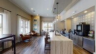 This single-story coffee bar in Gonzales, Louisiana, is thoughtfully designed to blend functionality with an inviting, modern aesthetic. The space centers around a striking marble-topped island with dramatic veining, fitted with a sink and surrounded by upholstered bar seating. Three glass globe pendant lights hang overhead, adding warmth and visual interest against the elevated ceiling with crown molding detail.

Behind the island, a dedicated beverage station is framed by a decorative patterned tile backsplash and outfitted with professional coffee and brewing equipment set against shiplap-style wall paneling. Built-in desk nooks with computer workstations flank the beverage area, providing practical workspace within the lounge environment.

Rich wood-look flooring extends throughout, grounding the neutral warm-toned walls and creating a cohesive, welcoming atmosphere. A relaxed seating area near the windows features leather lounge chairs and accent tables, bathed in natural light from multiple large windows dressed with white curtains. Additional high-top table seating positioned by the windows offers more casual spots to gather. Colorful framed artwork adorns the walls, adding a curated, polished finishing touch to the overall space.
