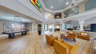 This generously sized single-floor space is designed as an open, multi-functional community gathering area with a refined yet welcoming aesthetic. Architecturally, the space is defined by its dramatically tall ceilings accented with a painted tray detail in a soft blue-grey hue, elaborate crown molding, and a series of white arched colonnades that elegantly divide functional zones while maintaining a sense of openness and flow throughout.

The main living and lounge area is anchored by a starburst chandelier suspended from the soaring ceiling and features clusters of amber leather seating arranged around a central coffee table atop a patterned area rug. Large floor-to-ceiling windows along one wall flood the space with natural light and offer views of lush outdoor greenery and a pool area beyond.

A dedicated dining zone includes a long communal wood table with cross-back chairs, while a separate recreation area accommodates a billiards table, bar-height seating, and vibrant wall art. A private office workspace features a built-in desk and arched transom window detail. Light wood-look flooring runs consistently throughout, unifying each zone. Lush tropical plants placed throughout enhance the bright, airy, resort-inspired ambiance.