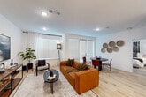 This thoughtfully designed single-floor residence combines modern aesthetics with practical functionality. The open-concept main area seamlessly integrates the living room, dining room, and kitchen, creating a bright and cohesive space. Large windows and a glass door flood the living area with natural light, complemented by light-toned hardwood flooring and recessed lighting throughout. The kitchen is outfitted with dark wood cabinetry, granite countertops, stainless steel appliances including a full-size refrigerator, range, and built-in microwave, alongside a double sink — offering a well-equipped cooking environment. A dedicated dining area features direct access to the outdoors via a glass door, while a built-in desk nook provides a convenient workspace. The bedroom is a serene retreat with plush carpeting, a ceiling fan, ample natural light from a large window with curtains, and generous closet space — four closets are distributed throughout the unit. The full bathroom includes a dual-sink vanity with granite countertops, dark wood cabinetry, a large framed mirror, and a soaking tub with shower. Additional utility and hallway spaces contribute to the residence's overall practicality and well-organized flow.