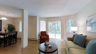 This two-floor space offers a comfortable and well-lit living environment defined by clean lines and a neutral color palette. The living area serves as the heart of the main level, featuring warm hardwood flooring that extends throughout the room and contributes to an inviting atmosphere. Large sliding glass doors flanked by additional windows flood the space with natural light while framing pleasant views of surrounding greenery, creating a seamless connection between the interior and the outdoors. A textured ceiling adds subtle visual interest overhead, while baseboard heating units are positioned along the walls, providing practical climate control. The layout is open enough to accommodate a comfortable seating arrangement, as evidenced by the combination of a sofa and accent chair, centered around a round wooden coffee table. Structural columns punctuate the space without interrupting the overall flow of the room. The light-toned walls complement the natural light, enhancing the airy feel throughout. The second floor adds additional living space, further extending the functionality of this property. Overall, the space balances practicality with a bright, relaxed ambiance suited for everyday living.