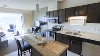 This single-floor space centers around a thoughtfully designed kitchen that balances functionality with style. The kitchen is anchored by a generously sized island with a granite countertop, offering ample prep space and casual seating, making it a natural gathering point. Tan placemats, glassware, and tableware displayed on the island highlight its dual role as a dining and entertaining surface.

Dark espresso cabinetry lines the walls, providing substantial storage while creating a bold contrast against the light-toned walls and granite surfaces. Stainless steel appliances, including an electric range, over-the-range microwave, and a double-basin sink, lend a modern, cohesive look to the space. A large overhead range hood serves as a subtle architectural focal point above the cooktop.

Natural light enters through a window near the entryway, brightening the open layout that connects the kitchen to an adjacent living area. A pendant light fixture above the island adds focused, ambient illumination. The living area, visible in the background, features a TV setup and side seating, reinforcing the open-concept flow between the kitchen and common living spaces.