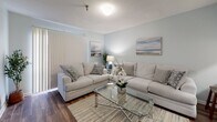 Located at 2633 Leaphart Rd in West Columbia, South Carolina, this single-floor residence presents a cohesive, light-filled layout designed for comfortable everyday living. Soft blue-grey walls and wood-look flooring run throughout the space, creating a calm, unified aesthetic from room to room.

The galley-style kitchen is efficiently organized, featuring white cabinetry with warm wood-toned trim on both sides, ample upper and lower storage, a double-basin sink, and white appliances including a coil-top range and full-size refrigerator. Neutral wall art and a light runner add a polished finishing touch.

The living area is generously proportioned, accommodating a sectional and sofa arrangement centered around a glass coffee table. Vertical blinds along a large window allow for natural light control, while the textured ceiling and wood-look flooring reinforce the cohesive interior palette.

The bedroom is a serene retreat, featuring the same wood-look flooring, soft walls, a wide closet with vertical blinds, and ample space for a full furniture arrangement. The bathroom includes a walk-in shower stall with grab bar, a curtained enclosure, and warm wood-look flooring, maintaining the clean, neutral character found throughout the residence.