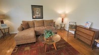This single-floor living space features a cozy and well-appointed living room with warm hardwood flooring that extends throughout the area, contributing to an inviting atmosphere. The room is anchored by a generously sized sectional sofa upholstered in a tan microfiber fabric, complemented by decorative throw pillows in rich earth tones. A round glass-top coffee table with wooden legs sits atop a bold, patterned area rug in deep reds and golds, adding visual interest and warmth to the space. A matching glass-top side table with a table lamp provides ambient lighting in the corner, accompanied by a woven accent chair that adds texture and character. Wall décor includes a framed botanical print in a dark wood frame, enhancing the classic, understated aesthetic. The neutral wall color creates a clean backdrop that highlights the warm tones of the furnishings and flooring. The overall ambiance is comfortable and approachable, with a cohesive color palette of browns, reds, and greens that ties the space together in a welcoming and functional arrangement suited for everyday living.