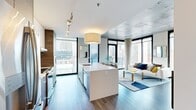 This contemporary urban residence offers two bedrooms and two bathrooms across a single thoughtfully designed floor. The open-concept living and kitchen area is an immediate highlight, featuring floor-to-ceiling windows that frame expansive city skyline views and flood the space with natural light. The kitchen is anchored by a sleek white island with an integrated sink, complemented by wood-toned cabinetry, stainless steel appliances, and track lighting overhead. Exposed concrete ceilings and structural columns lend an industrial character that contrasts beautifully with the clean, modern finishes throughout.

The living area connects seamlessly to a private balcony through large glass doors, offering unobstructed views of the surrounding cityscape, including a prominent domed landmark building. Both bedrooms continue the floor-to-ceiling window theme, providing abundant natural light and striking urban vistas. Exposed ductwork and concrete accent walls reinforce the industrial-modern aesthetic.

The primary bathroom is a refined retreat, featuring marble-style tile flooring, dual vessel sinks on a wood-framed vanity, a large wall mirror, and a glass-enclosed walk-in shower. The second bathroom offers a clean, minimalist design with a vessel sink and a shower-tub combination. Rich hardwood flooring runs throughout the main living areas, completing this polished, city-living experience.