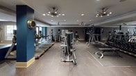 Located at 5940 Forest Park Rd in Dallas, Texas, this single-floor fitness center is a comprehensively equipped facility designed to support a full range of workout routines. The space is finished with a contemporary aesthetic, featuring a neutral and navy blue color palette, bold structural columns, and a combination of patterned carpet and hard flooring throughout.

The cardio zone is well-stocked with a row of Matrix treadmills and additional cardio machines, including ellipticals and upright bikes, positioned for ease of use. Wall-mounted televisions are integrated into the space, keeping users entertained during workouts. The strength training area features multiple selectorized cable and resistance machines suited for a variety of muscle groups, complemented by a dedicated free weights section with a full rack of dumbbells and an adjustable bench.

Floor-to-ceiling mirrors line key walls, visually expanding the space and allowing users to monitor their form. Recessed lighting and ceiling fan fixtures provide consistent illumination and airflow throughout. A medicine ball rack adds functional training options to the layout. Natural light filters in through windows, offering views of the surrounding outdoor environment. The overall ambiance is modern, purposeful, and well-maintained, making this fitness center a highly functional space for daily exercise.