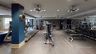 Located at 5940 Forest Park Rd in Dallas, Texas, this single-floor fitness center is a comprehensively equipped facility designed to support a full range of workout routines. The space is finished with a contemporary aesthetic, featuring a neutral and navy blue color palette, bold structural columns, and a combination of patterned carpet and hard flooring throughout.

The cardio zone is well-stocked with a row of Matrix treadmills and additional cardio machines, including ellipticals and upright bikes, positioned for ease of use. Wall-mounted televisions are integrated into the space, keeping users entertained during workouts. The strength training area features multiple selectorized cable and resistance machines suited for a variety of muscle groups, complemented by a dedicated free weights section with a full rack of dumbbells and an adjustable bench.

Floor-to-ceiling mirrors line key walls, visually expanding the space and allowing users to monitor their form. Recessed lighting and ceiling fan fixtures provide consistent illumination and airflow throughout. A medicine ball rack adds functional training options to the layout. Natural light filters in through windows, offering views of the surrounding outdoor environment. The overall ambiance is modern, purposeful, and well-maintained, making this fitness center a highly functional space for daily exercise.