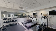 Located at 3528 Gentian Blvd in Columbus, Georgia, this fitness center is a well-equipped, single-room facility designed to accommodate a full range of exercise routines. The space features a drop-panel ceiling with recessed lighting that provides even, bright illumination throughout. Light wood-look flooring runs across the open area, lending a clean and modern aesthetic to the interior.

The room is lined with large wall mirrors that visually expand the space and allow users to monitor their form during workouts. Cardio equipment is a central focus, with multiple treadmills and an elliptical machine arranged along one wall, accompanied by a wall-mounted flat-screen television for entertainment during exercise. Protective rubber floor mats are positioned beneath the equipment for safety and durability.

A dedicated free-weight zone features a multi-tier dumbbell rack stocked with a range of weights, alongside an adjustable workout bench and exercise mats, providing ample options for strength training. A water dispenser and waste receptacle are conveniently placed near the entrance area.

The facility includes an ADA-compliant restroom and a glass-panel exterior door that opens to an outdoor area, allowing for natural light and ventilation. Informational signage is posted near the entrance. The overall design is clean, functional, and efficiently organized to maximize usability within its compact footprint.