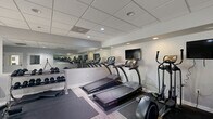Located at 3528 Gentian Blvd in Columbus, Georgia, this fitness center is a well-equipped, single-room facility designed to accommodate a full range of exercise routines. The space features a drop-panel ceiling with recessed lighting that provides even, bright illumination throughout. Light wood-look flooring runs across the open area, lending a clean and modern aesthetic to the interior.

The room is lined with large wall mirrors that visually expand the space and allow users to monitor their form during workouts. Cardio equipment is a central focus, with multiple treadmills and an elliptical machine arranged along one wall, accompanied by a wall-mounted flat-screen television for entertainment during exercise. Protective rubber floor mats are positioned beneath the equipment for safety and durability.

A dedicated free-weight zone features a multi-tier dumbbell rack stocked with a range of weights, alongside an adjustable workout bench and exercise mats, providing ample options for strength training. A water dispenser and waste receptacle are conveniently placed near the entrance area.

The facility includes an ADA-compliant restroom and a glass-panel exterior door that opens to an outdoor area, allowing for natural light and ventilation. Informational signage is posted near the entrance. The overall design is clean, functional, and efficiently organized to maximize usability within its compact footprint.