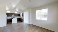 This single-story space showcases an open-concept design that seamlessly connects the living area and kitchen. The flooring throughout is consistent hardwood-style planking in a warm, medium-toned finish, lending continuity and a clean aesthetic across the space.

The living area benefits from generous natural light, courtesy of a large window that frames an outdoor view of neighboring structures and mature trees. A vertical blind panel provides privacy control near what appears to be a sliding door, offering potential outdoor access.

The kitchen is defined by dark espresso-toned cabinetry arranged in an L-shaped configuration, creating a strong visual contrast against the neutral, light-toned walls. Stainless steel appliances, including a microwave and range, are integrated into the cabinetry. An additional clerestory-style window above the kitchen cabinets introduces supplemental natural light into the cooking area.

The ceiling features a subtle vaulted section over the kitchen, adding vertical dimension to the space. Recessed lighting and a ceiling fan mount are also present. The overall ambiance is clean, modern, and functional, with a neutral palette that offers versatility for various interior styles.