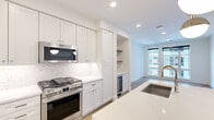 This well-appointed one-bedroom, one-bathroom residence offers a refined living experience across a single floor. The open-concept layout flows naturally from the kitchen into the living area, bathed in natural light from large windows with exterior views.

The kitchen is a standout feature, showcasing floor-to-ceiling white shaker cabinetry, a striking two-tone island with navy blue lower cabinets and a crisp white quartz countertop, and a full suite of stainless steel appliances including a professional-grade gas range, built-in microwave, French door refrigerator, dishwasher, and a built-in wine cooler. A herringbone marble backsplash and open wood shelving add warmth and texture. Light hardwood flooring carries throughout the main living areas.

The living room provides an airy, versatile space with large windows that draw in abundant natural light. The bedroom features plush carpeting, a ceiling fan, and a window overlooking the surrounding neighborhood.

The bathroom is elegantly finished with full marble-look tile on both the floors and walls, a frameless glass walk-in shower with a built-in niche and bench seat, and chrome fixtures. An adjacent walk-in closet with built-in shelving and hanging storage adds practical functionality to the private quarters.