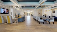 Located at 1209 Westwood Ave in Richmond, Virginia, this single-floor community clubhouse presents a thoughtfully designed interior that balances elegance with functionality. The welcoming entrance lobby features a striking herringbone-patterned floor, a contemporary multi-globe chandelier with brass accents, and a reception area framed by large glass panels that draw in natural light and provide views of the surrounding exterior.

The main gathering hall is anchored by coffered ceilings painted in a rich teal-blue, offset by crisp white trim and warm wood-look plank flooring. This expansive area is divided into distinct zones: a comfortable lounge seating area furnished with curved sofas and accent chairs, and a formal dining zone centered around a long dark conference-style table surrounded by upholstered chairs, illuminated by geometric gold pendant fixtures. Large windows and glass doors open to the outdoor surroundings, ensuring the interior remains bright throughout the day.

A well-appointed kitchenette serves the space, featuring dark cabinetry with gold hardware, white quartz countertops, stainless steel appliances, a wall-mounted television, and a textured geometric tile backsplash. The overall ambiance is polished and contemporary, making this a versatile space suited for both social gatherings and community events.