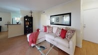 This single-floor residence offers a thoughtfully designed layout that balances functionality with comfort. The kitchen is outfitted with white cabinetry, dark granite-style countertops, and white appliances including a dishwasher and range, all set atop light tile flooring that adds brightness to the space. Crown molding details carry throughout the home, lending a polished, cohesive aesthetic. The living room features warm hardwood flooring, crown molding, and a wall-mounted television, creating an inviting gathering space filled with natural light. Adjacent to the living area, the dining space continues the hardwood flooring and benefits from a large window dressed with sheer curtains, adding an airy, open feel. The bedrooms are carpeted for comfort, each offering closet storage — including a walk-in closet visible in the primary bedroom — and windows that allow natural light to fill the rooms. The bathrooms are cleanly finished with white tile throughout; one features a soaking tub with a shower combination, while the other offers a standalone walk-in shower. Overall, the space presents a clean, neutral palette with warm wood accents that create a welcoming and practical living environment.