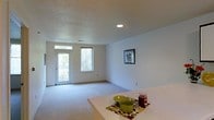 This single-floor space presents a clean, functional layout centered around an open living area that feels welcoming and practical. The living room is finished with wall-to-wall carpeting in a neutral tone, contributing to a comfortable and quiet atmosphere. White walls provide a bright, versatile backdrop throughout the space, while recessed ceiling lighting supplements the natural light that enters through multiple windows. The textured popcorn ceiling adds a classic residential character to the room.

Natural light is a notable feature of the living area, with a glass-paneled entry door flanked by a window, allowing generous daylight to fill the space and offering a view of surrounding greenery. The open sightlines between the living area and what appears to be a dining or kitchen counter area suggest a connected, flowing floor plan well-suited for everyday living and casual entertaining. A framed piece of wall art adds a touch of personality to the otherwise understated decor. Overall, the space offers a straightforward, move-in-ready environment with a neutral palette that lends itself easily to personalization.